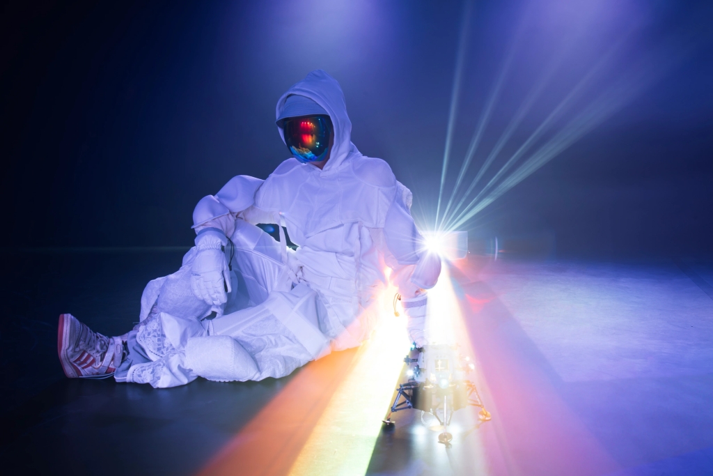 A Person in a white spacesuit wearing the white hood over the head and a shiny, reflective facecoverin black, red and blue is sitting comfortably on the ground. In front of the person there is a lego spaceship in grey, black with lights in blue. The person and the spaceship are being lit by a light coming from behind like a sunray. 